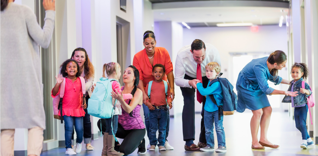 School staff and parents greeting young students during a school activity.